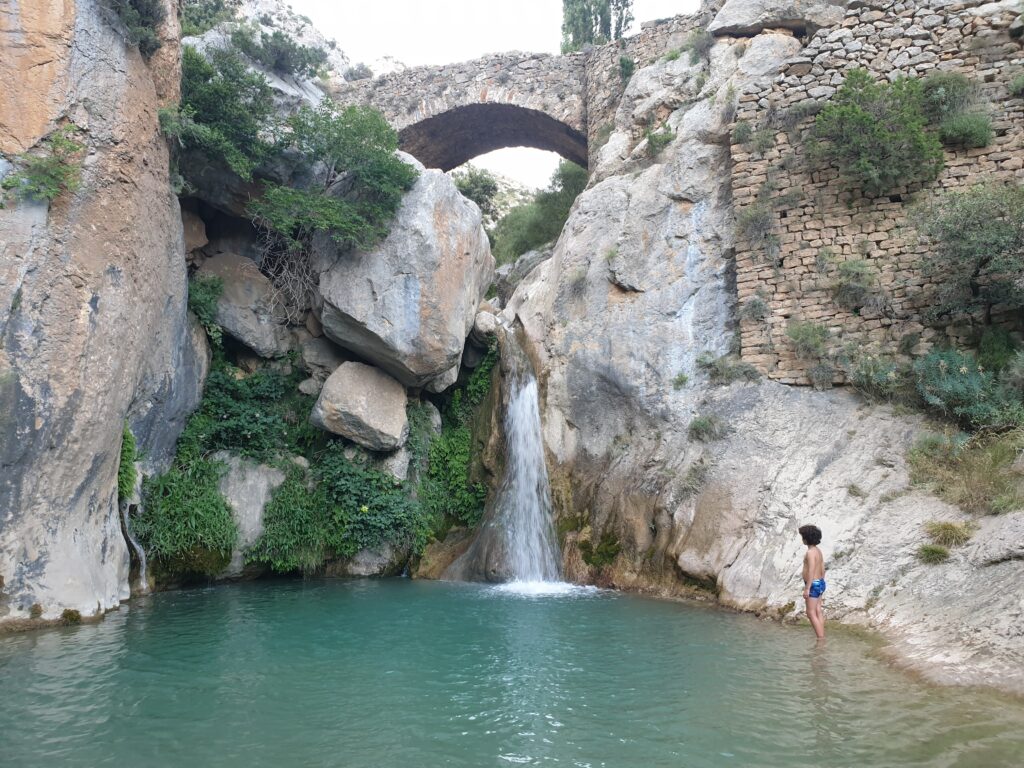 iño junto a la poza de la cascada de Bóixols en verano con el puente medieval al fondo